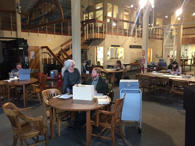 View of the reading room in the Maritime Research Center showing researchers, NPS uniformed staff as well as visitors, at tables consulting archives, books, and audio materials, Reference Librarian Gina Bardi at the Reference Desk in the background