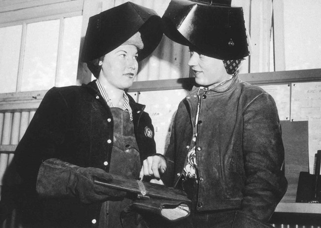 black and white image of two women welders standing together wearing welders' helmets with the face visor up