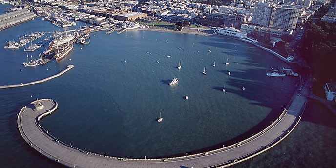 An aerial view of the park showing the curving Municipal Pier enclosing Aquatic Park cove. Hyde Street Pier and the fleet of historic vessels is on the left.