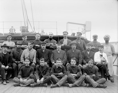 Twenty one men seated on the deck of a sailing ship for a formal pose of the ship's crew..