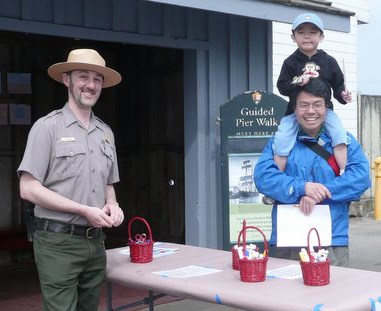A park ranger standing next to an adult carrying a child on his shoulders.