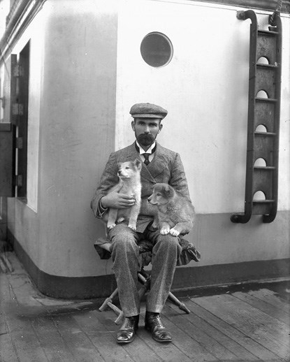 A black and white photo taken in 1905 of a man sitting on a stool onthe deck of a ship with two puppies on his lap.