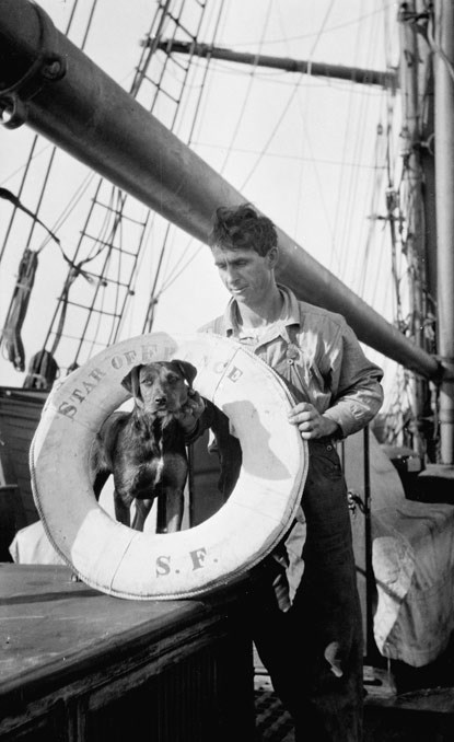 A man standing on the deck of a ship holding a ship’s ring around his black-colored dog.