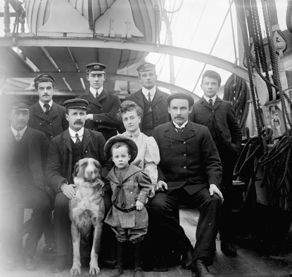 Family members, crew and large dog posing for a formal portrait on the deck of a large sailing ship.