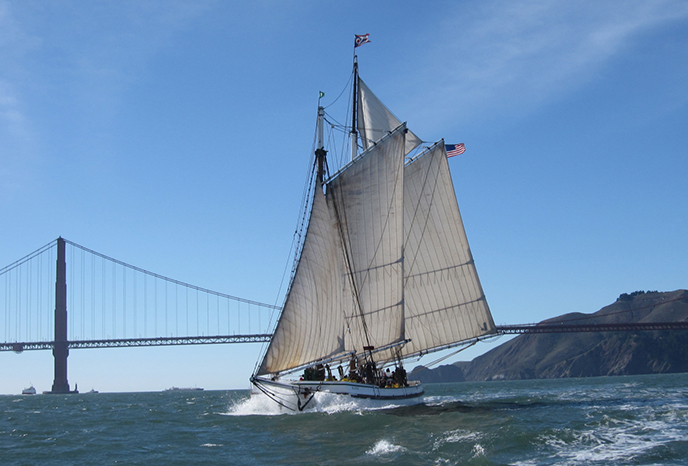 1891 flat-bottomed schooner on San Francisco Bay with her sails raised and the Golden Gate Bridge in the background