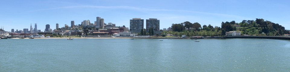 Looking across the calm Aquatic Park Cove water to the park’s Maritime Museum, with the San Francisco city skyline in the background.