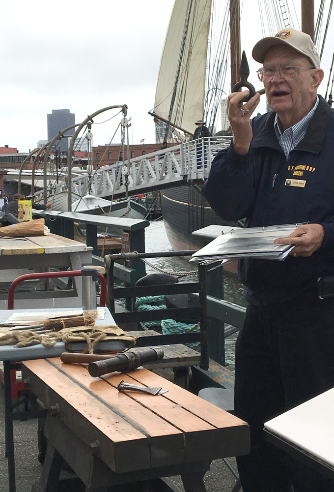 A man wearing a jacket with a docent badge, standing behind a table made of deck planks, holds up a traditional caulking tool.