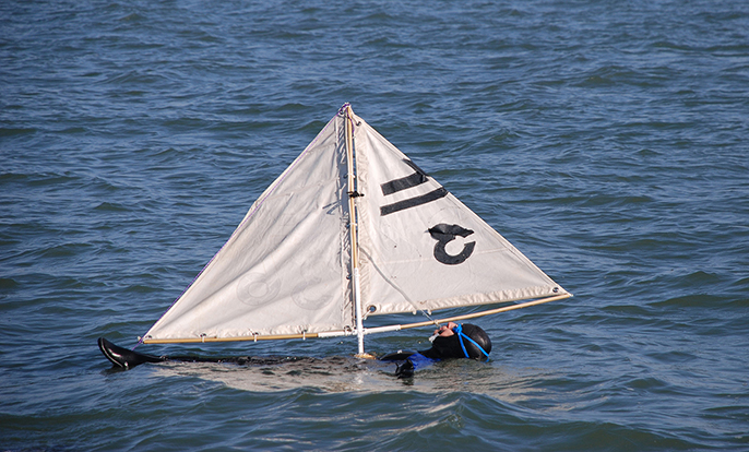 A man in a black wet suit floating in the water on his back on a surfboard with a mast and sails above him.