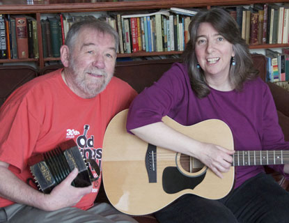 A man and woman seated next to each other and the man is holding a concertina and the woman is holding a guitar.