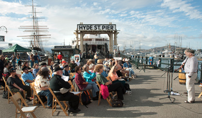 A group of people seated on Hyde Street Pier listening to two male performers singing.