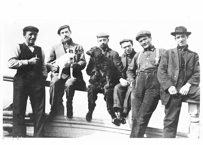 Historical photo of a group of sailors leaning against and sitting on railing. Two men in the middle are holding dogs on their laps.