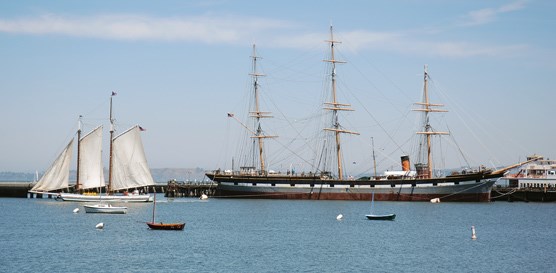 A view across a cove with a scow schooner with the sails raised and a 19th century sailing ship moored to a pier.