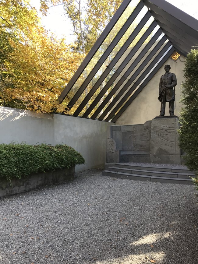 A bronze statue of a uniformed man stands on top of a bluestone pedestal with two women, words, and an exedra carved onto it.