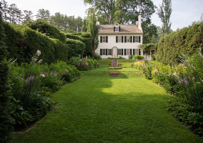 small lawn surrounded by flowers and hedges with large white building in background