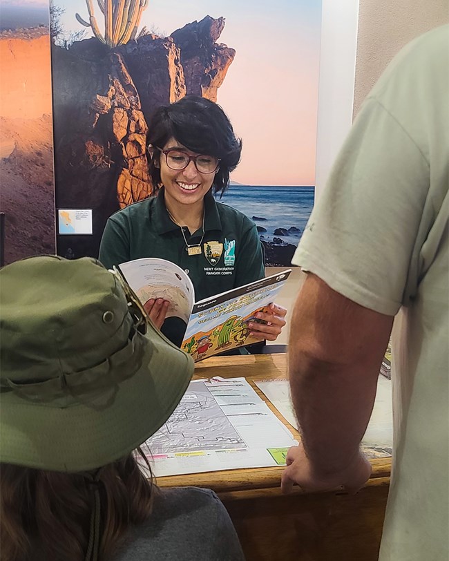 A young intern ranger sits at a desk reading a junior ranger book, with a father and small child standing on the opposite side of the desk.
