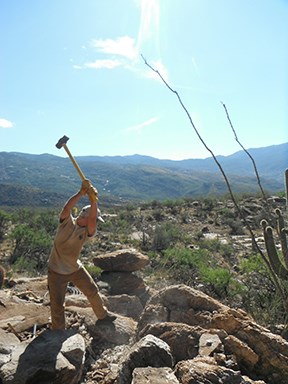 A trail crew member is up on the mountain swinging a tool.
