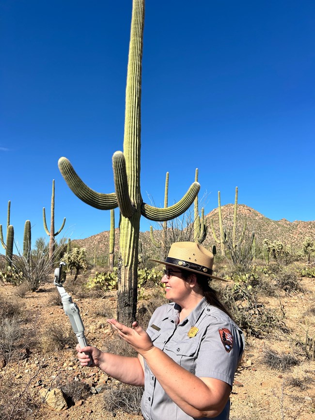 ranger stands in desert landscape, smiling and gesturing at a phone on a selfie stick