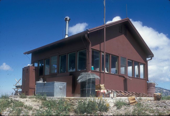 A small red shack takes up the majority of the photo, placed on a concrete slab. Green tinted windows wrap around the entire building. Barrels and storage crates pile around the building.
