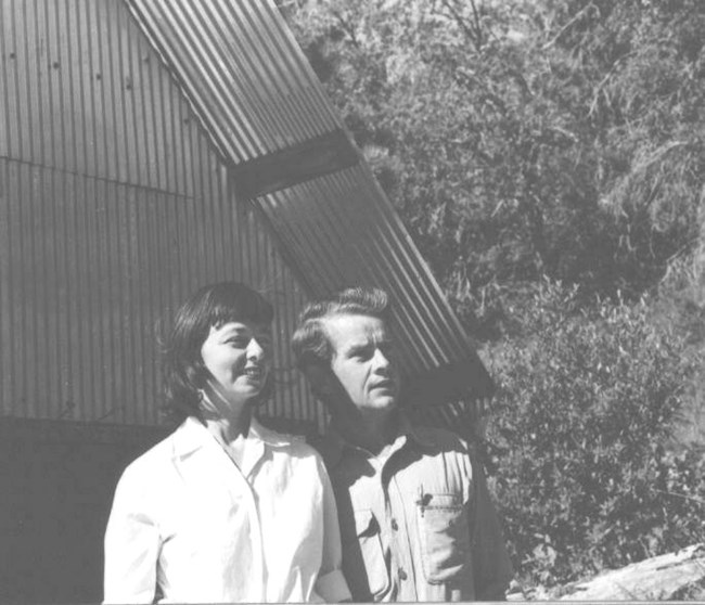 An old faded, black and white photo. A married couple stands in front of a metal shed, looking off to the right. Behind the shed is dense tree cover.