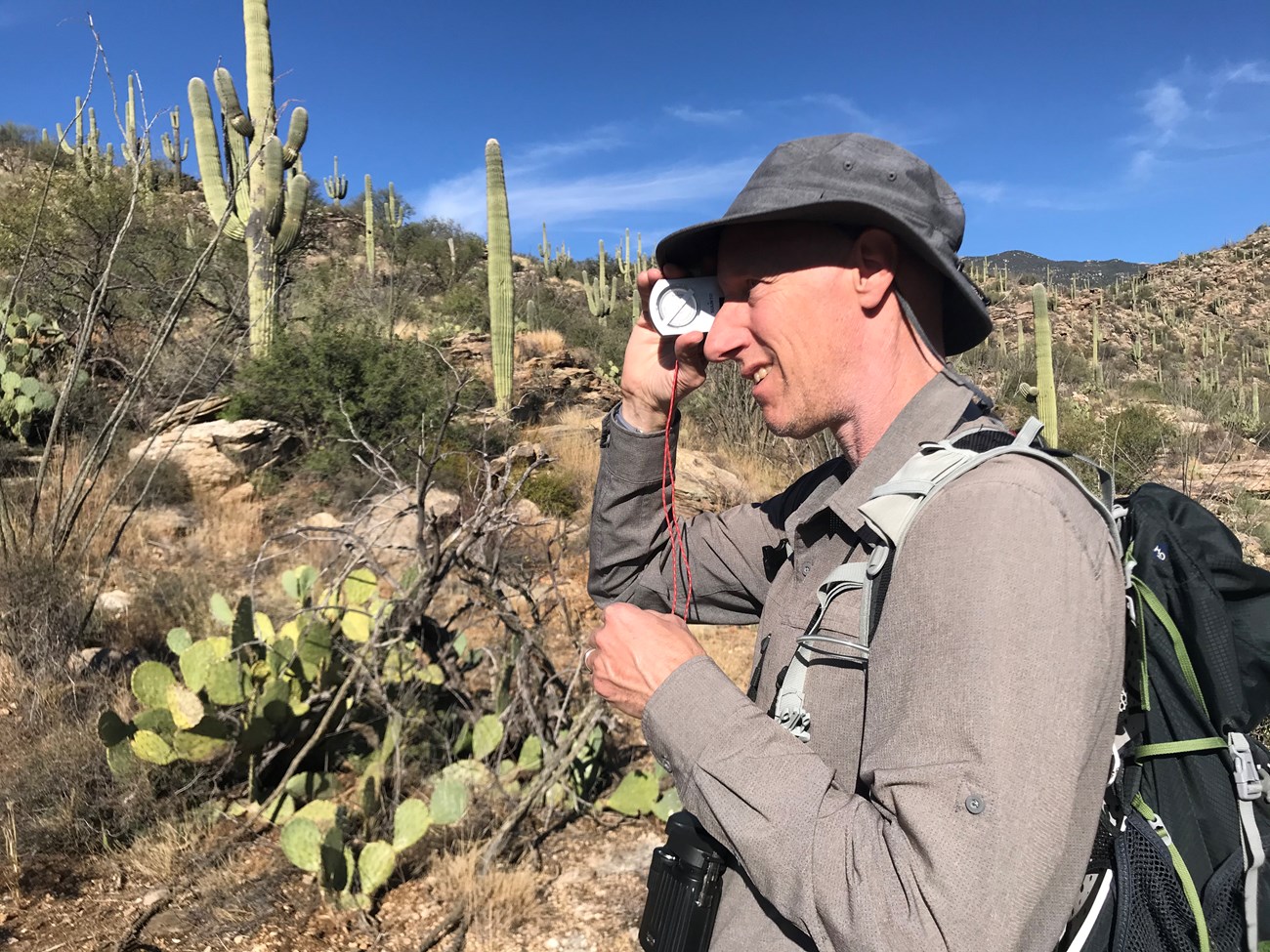 A side profile of a man wearing a grey hat and shirt. He is looking through a clinometer to find the height of a saguaro in front of him