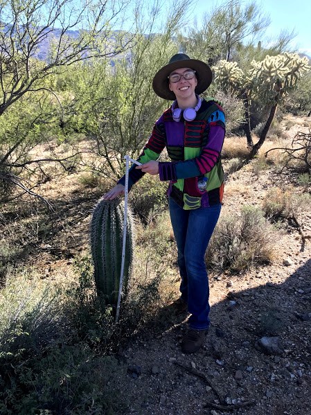 A female student standing and smiling next to a short saguaro. She is also holding a white folding ruler.