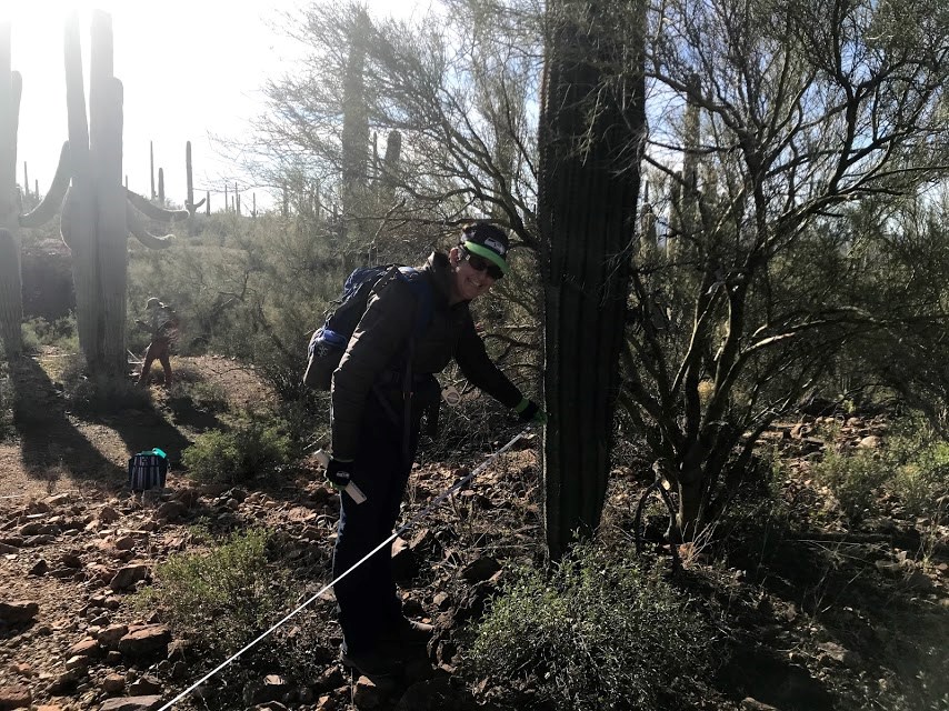 A woman smiling. She is holding the end of a tape measure next to the base of a saguaro.