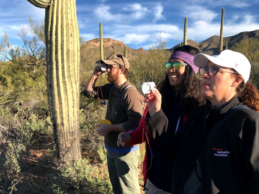 A man using a clinometer. Next to him are two women. One of them is teaching the other on how to use a clinometer.