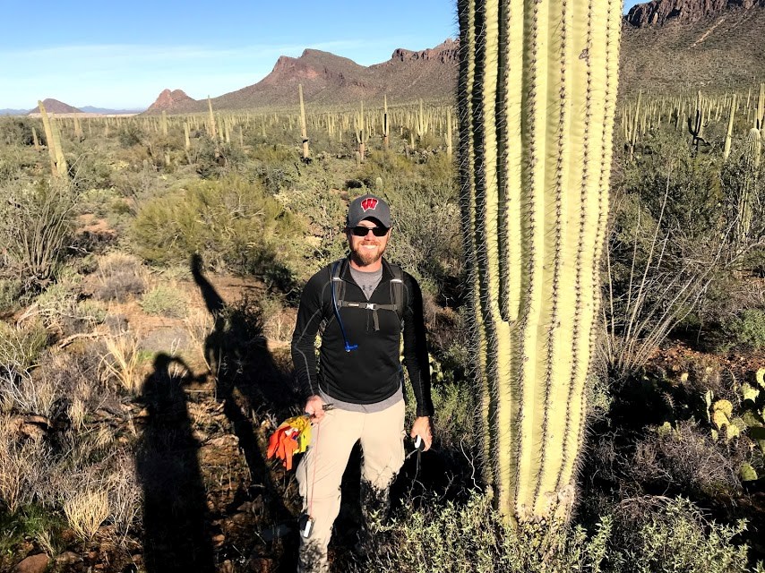 A man smiling to the camera. He is next to a big saguaro.
