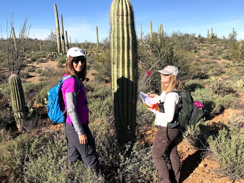 Two women smiling. In the middle of them is a tall saguaro with a red flag.