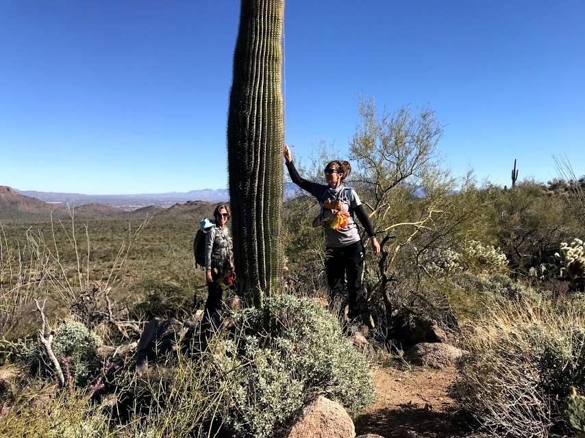 Two women on the plot smiling. One of them is putting a flag through the spines of a saguaro.
