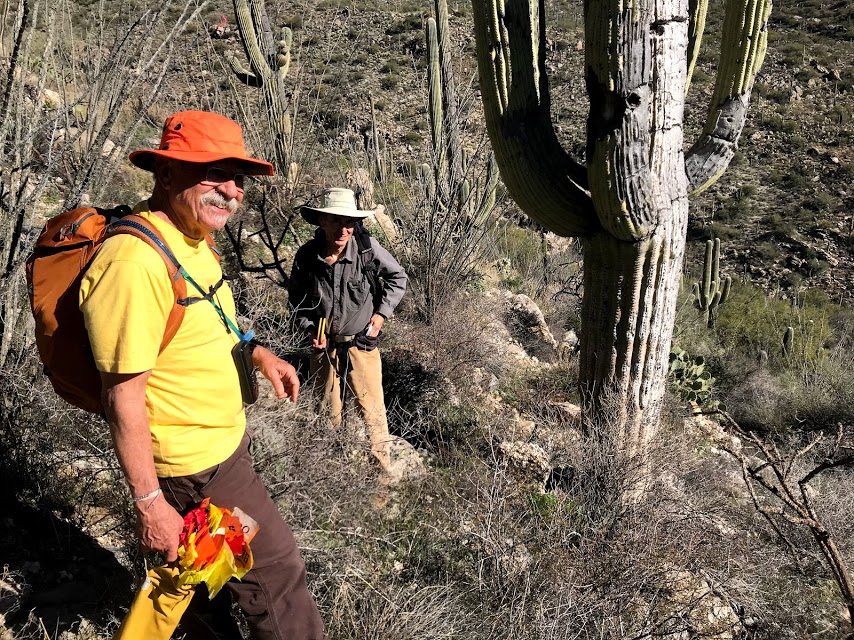 Two people smiling. One of them is holding a bag of flags while the other is holding a roll of measuring tape.