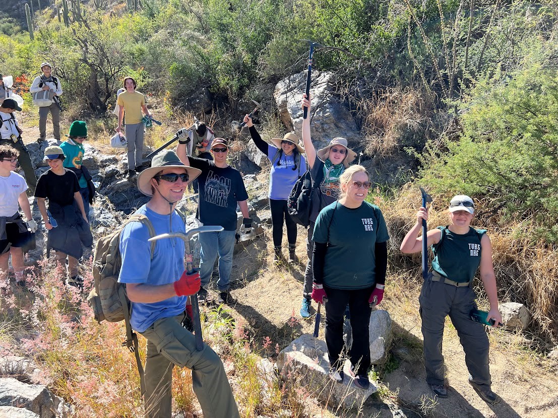 Horizontal image. A group of over a dozen volunteers stand along a dry riverbed, lined on either side with dense vegetation. They are wearing rough outdoor gear; thick cargo pants, gloves, etc. Many are carrying digging and pruning tools.