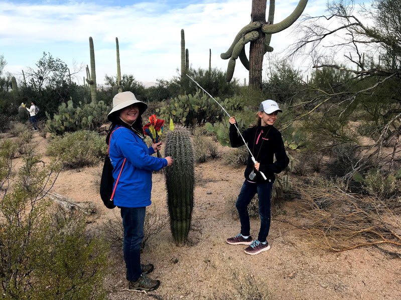 Two female volunteers holding flags and a meter stick near a small saguaro