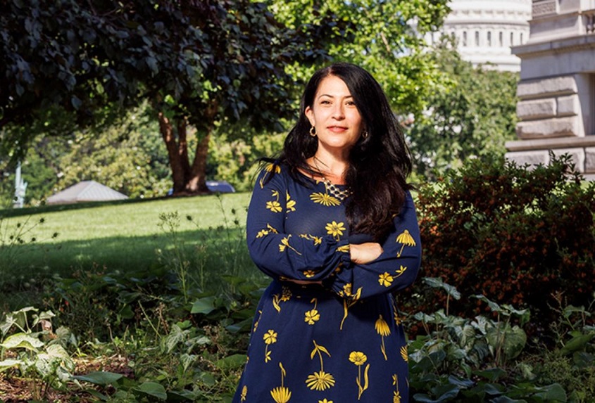 Ada_Limon_LOC A woman in a flowered dress stands in front of a city park with arms crossedpark with a