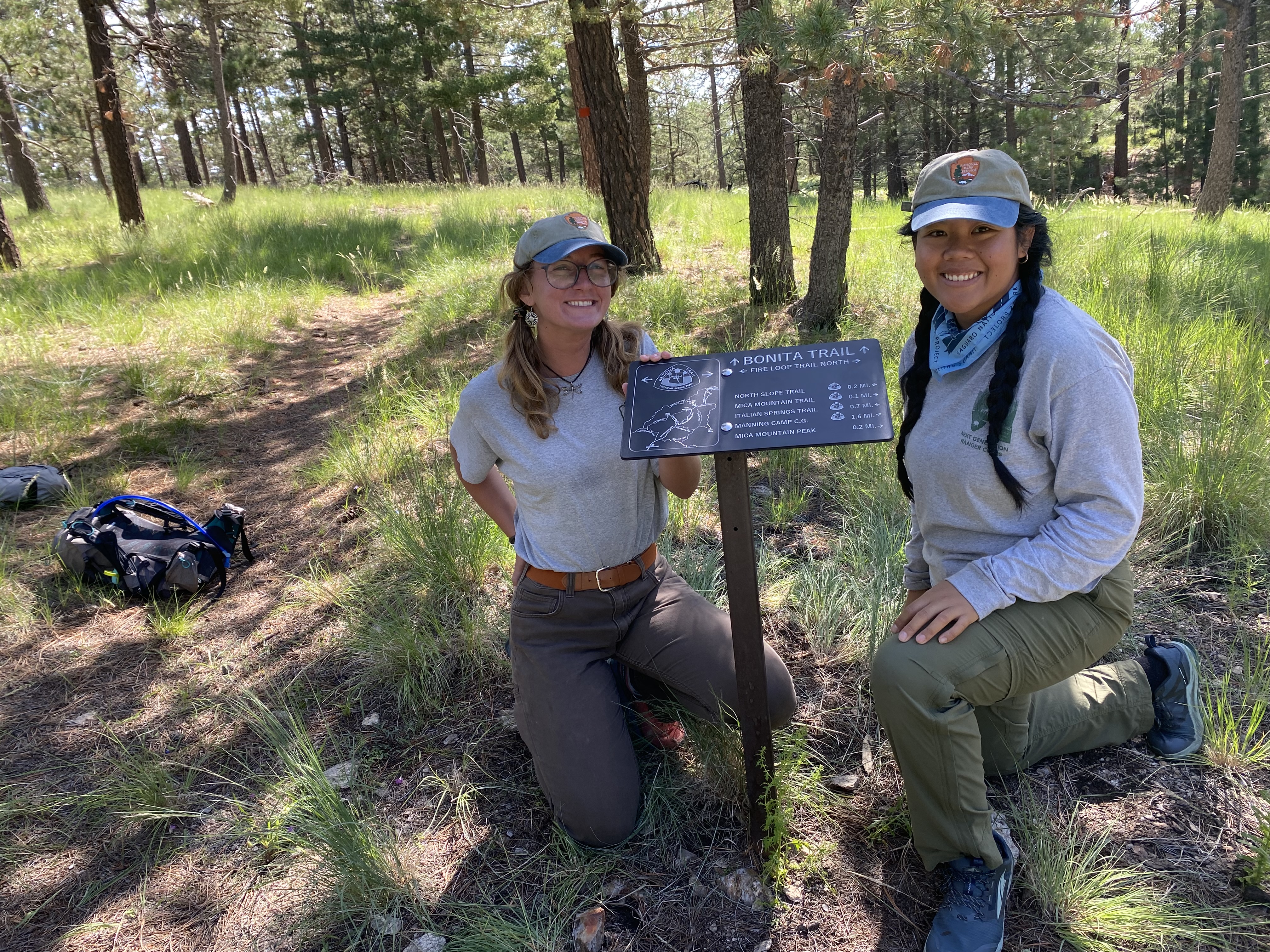 In the right of the image, two ranger interns kneel next to a small metal trail sign. Both wearing green pants, grey long sleeve shirts, and brown hats. To the left a trail winds around through a tree filled forest.