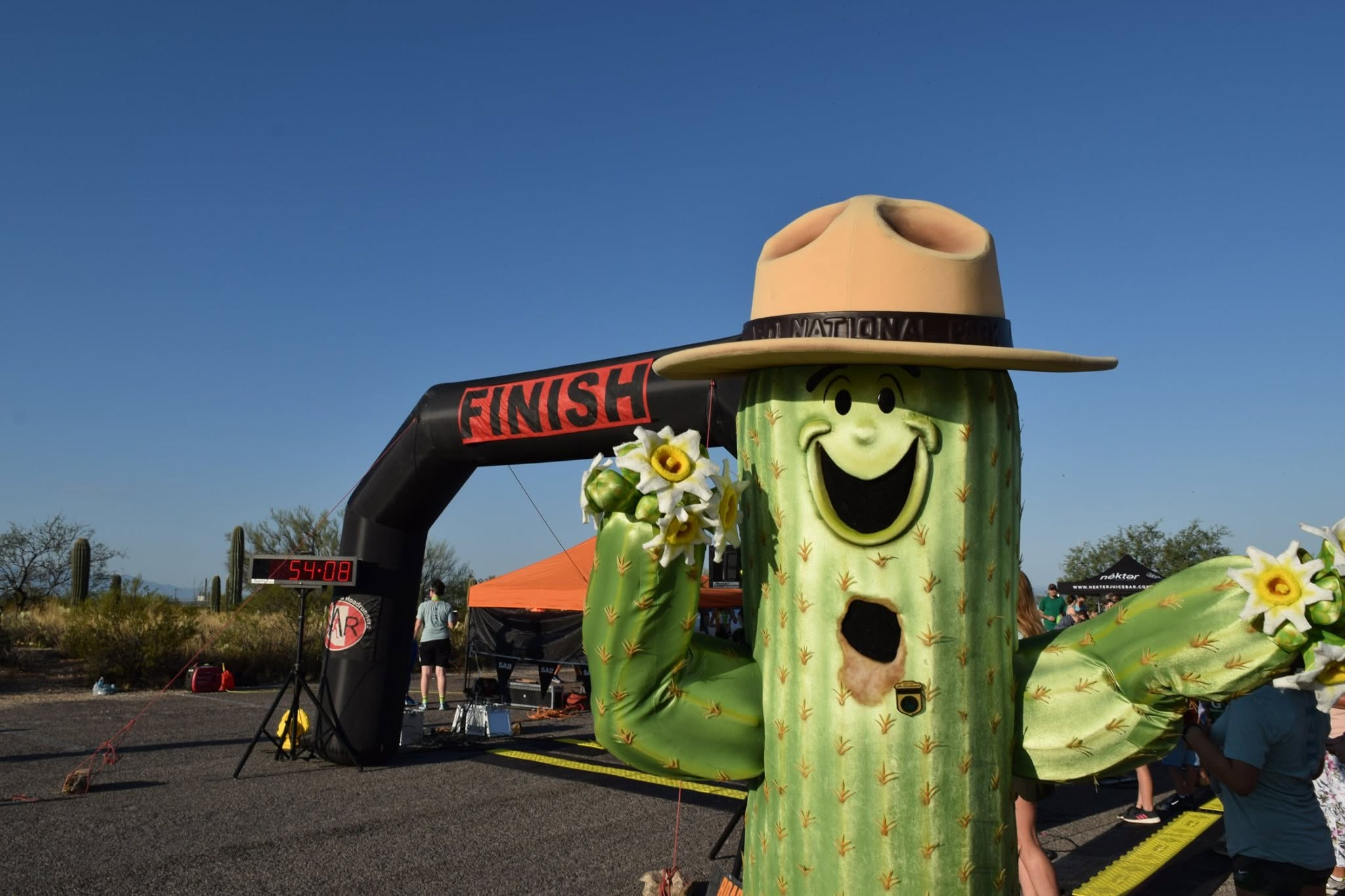 A happy Sunny the Saguaro mascot stands right of center, waving both arms. Behind him is a black inflatable arch reading "FINISH" with people going through.