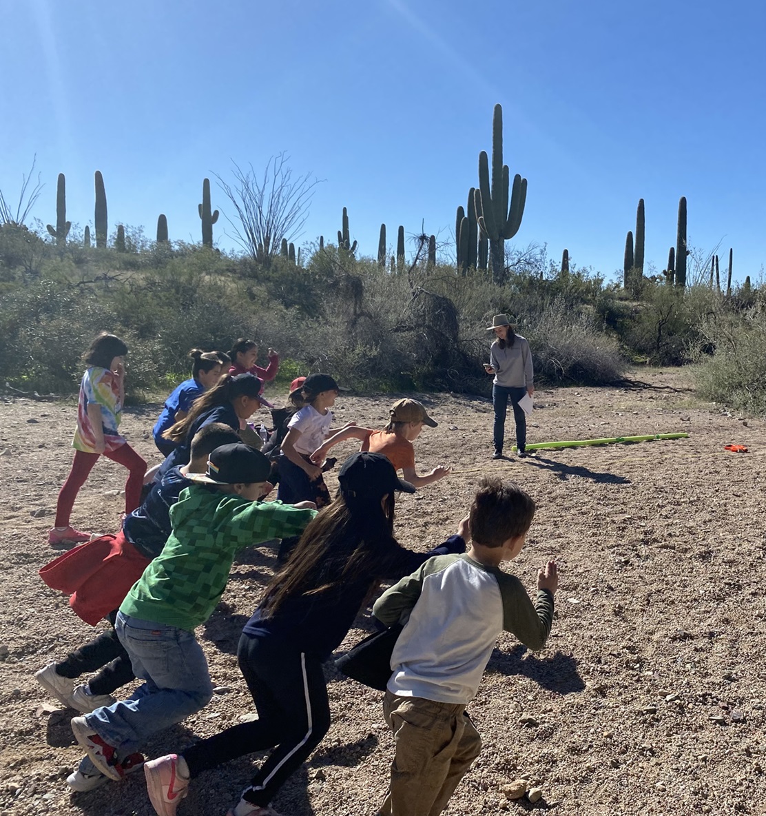 Ten kids sprint down a sandy wash as a ranger looks at a stopwatch beside them