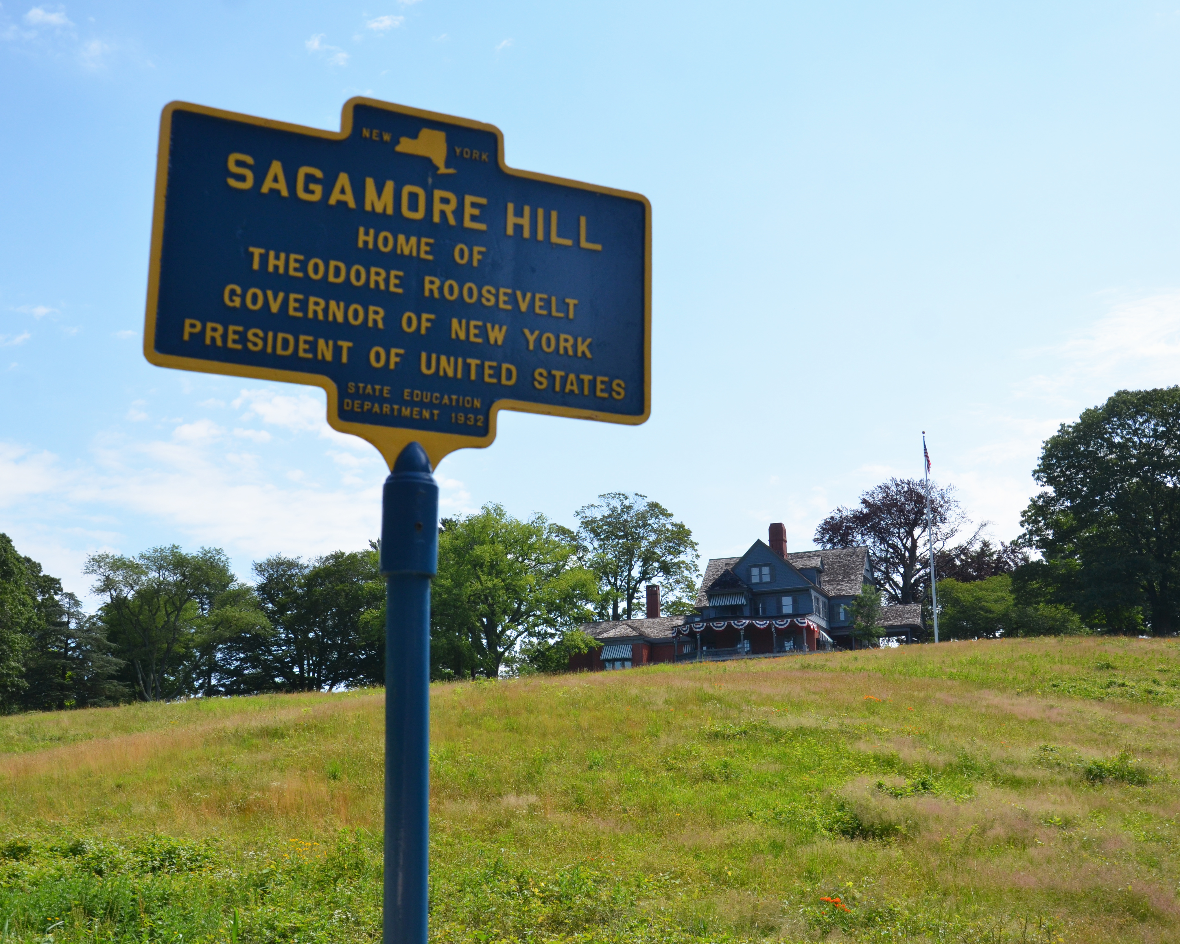 A blue sign reading, "Sagamore Hill, Home of Theodore Roosevelt, Governor of New York, President of United States" with the historic home atop a hill in the background.