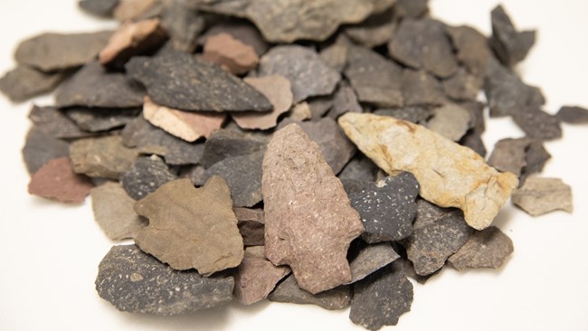 A variety of differently sized, shaped and colored chips of stone in a pile with five projectile points on top.