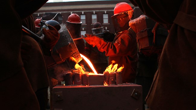 Several people wearing hard hats and facial shields surround a metal block. One man is pouring glowing molten metal onto block.