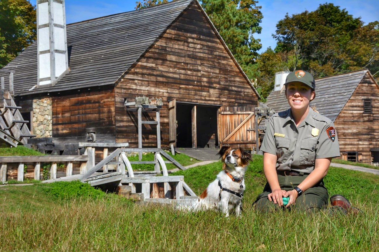park ranger sits in grass next to a small brown and white dog in front of two wood-sided buildings