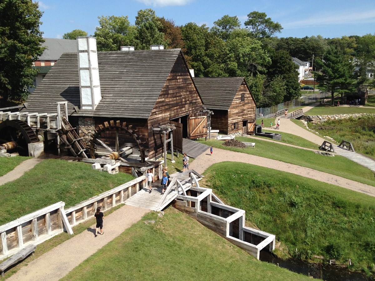 Visitors walk on paths around large wood-sided buildings with white chimneys and waterwheels.