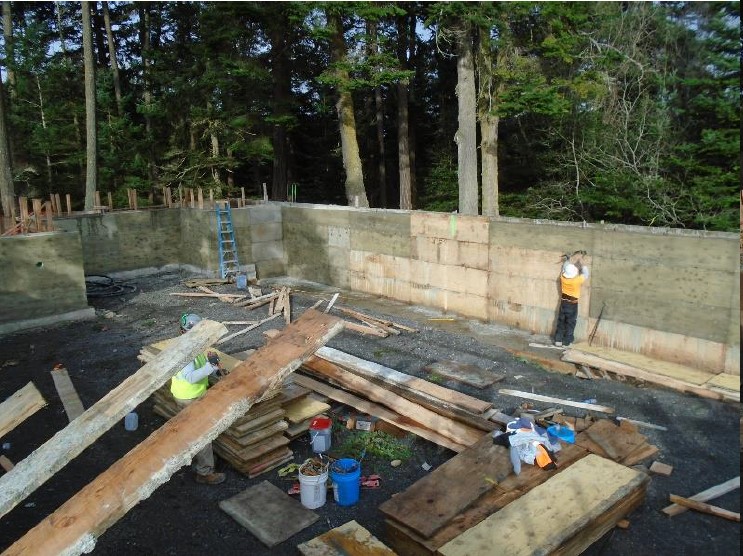 Stacks of plywood and wood boards are stacked in the foreground while a construction worker works on removing plywood sheets from the dried cement foundation walls