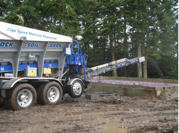 A construction truck with large truck bed full of gravel uses a conveyor belt to distribute gravel into the newly construction building foundation.