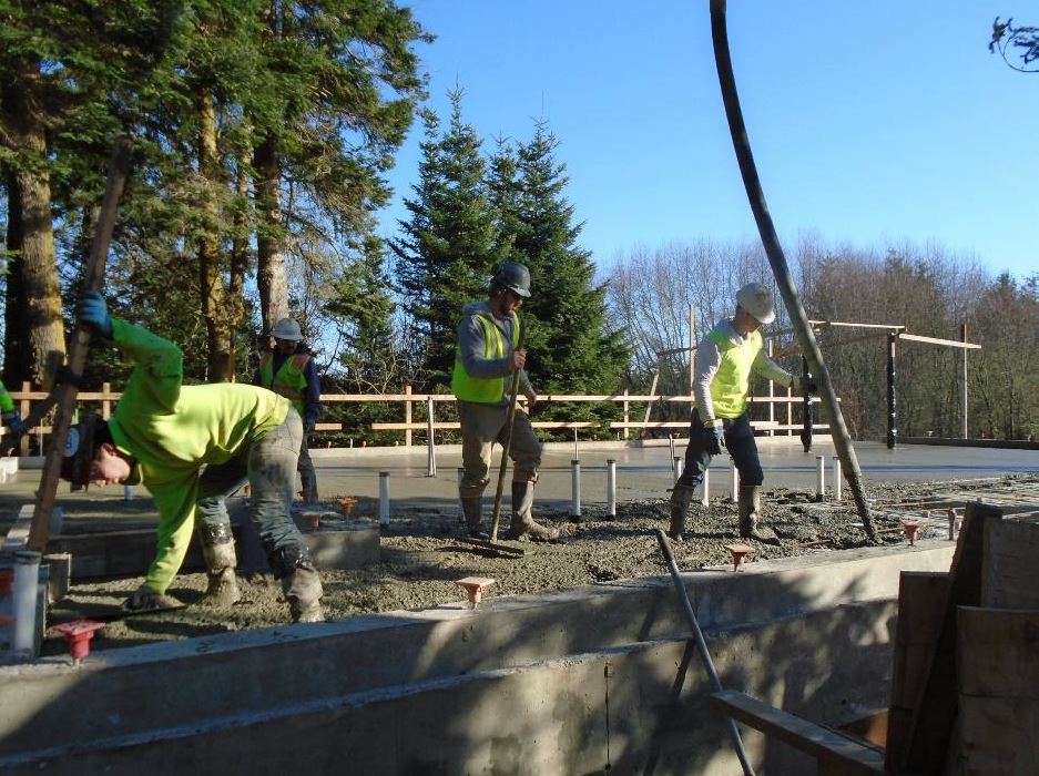 Four workers work to distribute cement with a hose and rakes throughout the foundation area