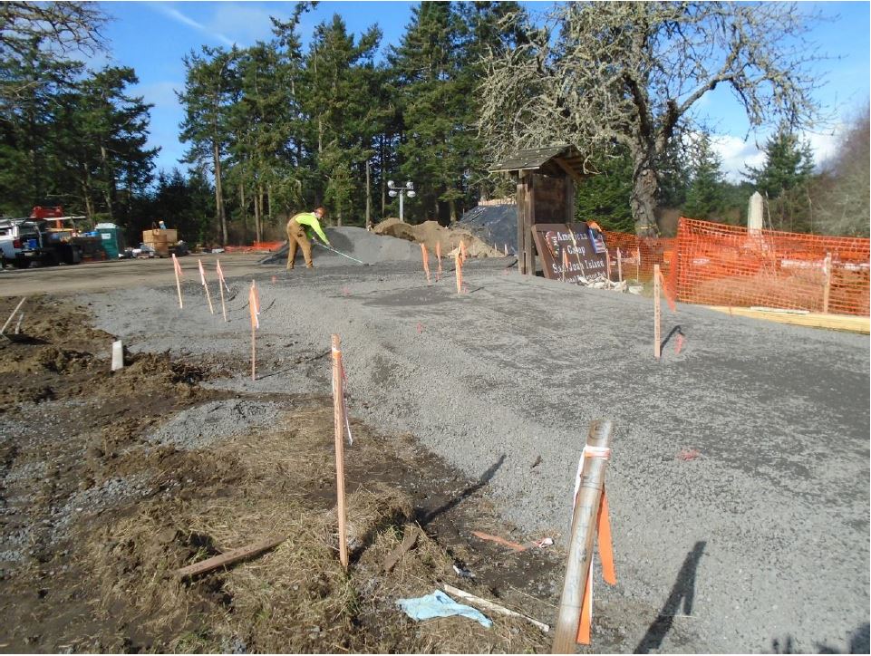 Survey markers mark the location of edge and center of the pedestrian pathway with gravel built up to form the pathway. A construction worker works in the background raking out the gravel for the path.