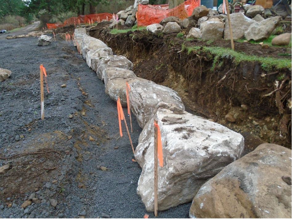 Large boulders are placed on gravel following survey markers along an earthen wall