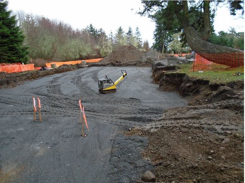 compacted sub-base aggregate fills out the newly excavated area where the visitor center will built. Surveyed stakes are in the foreground next to a compactor.