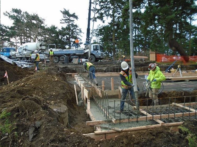 A construction worker guides a cement hose along building foundation footing framing in the foreground with a cement truck and crane in the background