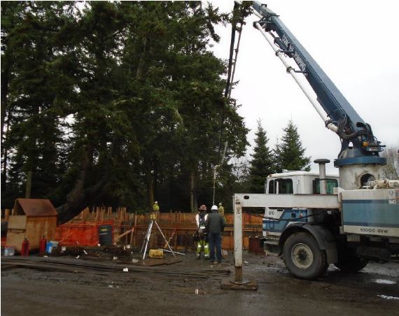 Cement truck in the foreground with a crane-like arm helps construction workers guide cement by a boom pump to the wooden foundation formwork.